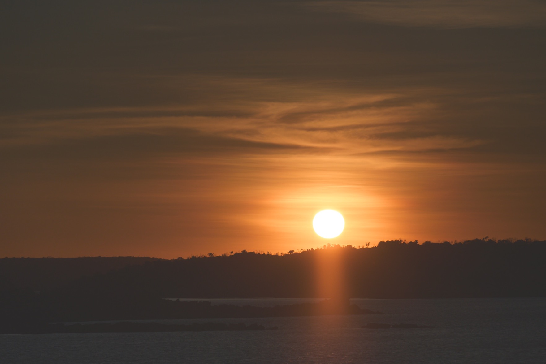 The sun breaking over the headlands, with a few clouds above and the water below.
